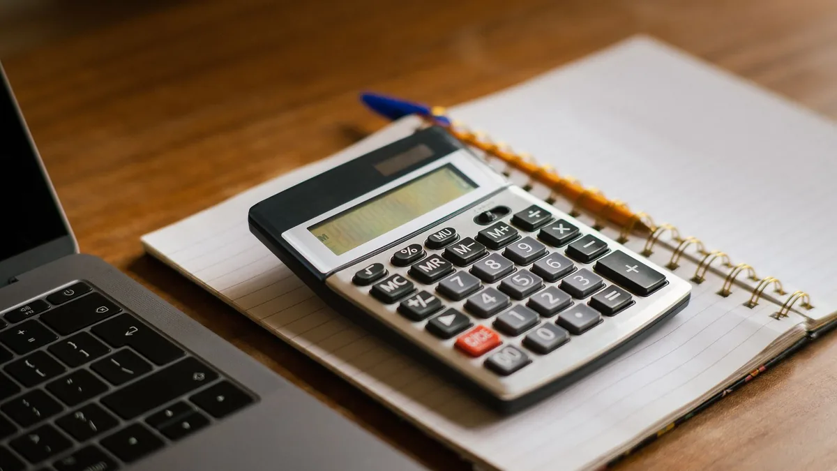 A calculator resting on a notepad beside a laptop on a wooden desk