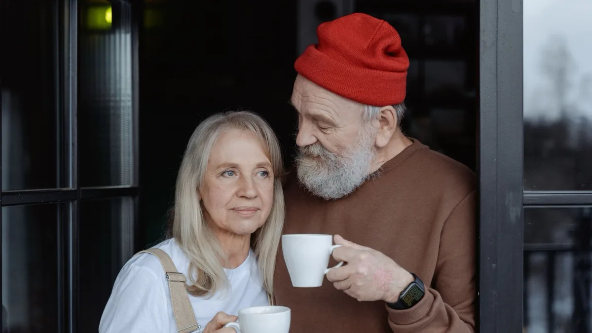An older couple standing together in a doorway holding coffee mugs, the man in a red knit cap looking toward the woman