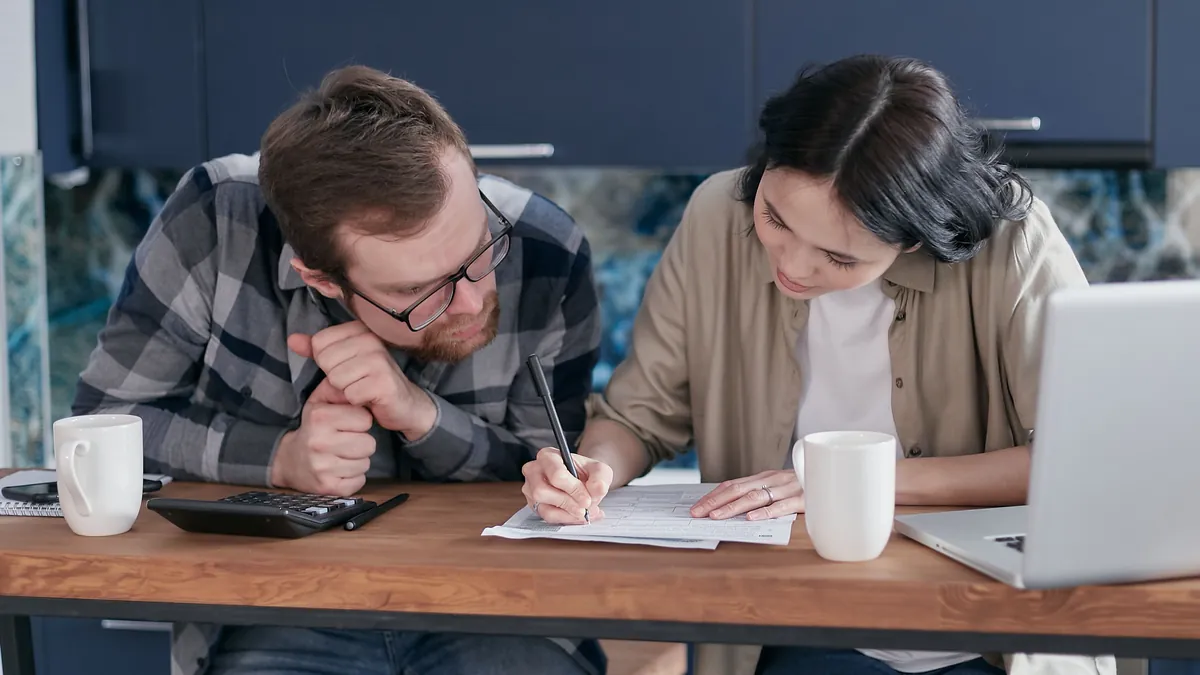 Two people at a kitchen table filling out a paper form together, a laptop and calculator beside them